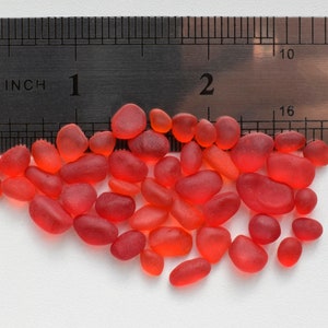 Peut inclure: Gros plan sur un tas de petits galets de verre lisses et rouges. Les galets sont éparpillés sur une surface blanche à côté d'une règle graduée en pouces.