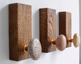May include: Three wooden wall hooks with stone knobs. The hooks are made of dark brown wood and the knobs are made of different colored stones. The hooks are mounted on a white wall.