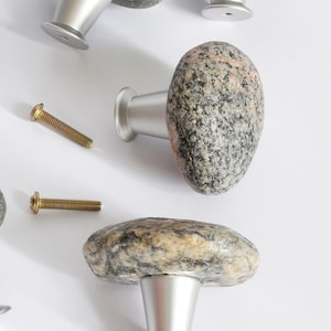 Pode incluir: Um conjunto de sete puxadores de armário de pedra com bases prateadas e parafusos de montagem. Os puxadores são feitos de pedras naturais em tons de cinza, castanho e rosa, oferecendo uma estética rústica. Para móveis e decoração.