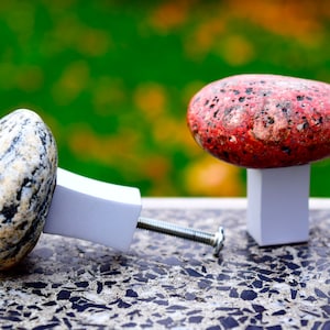 May include: Two stone cabinet knobs with white bases. One knob is grey and white with a natural stone finish. The other knob is red with a speckled finish.