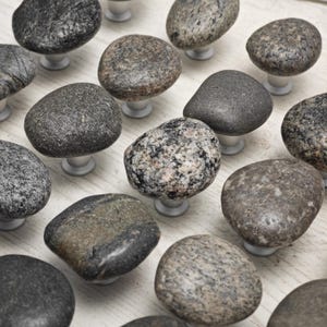 Pode incluir: Uma coleção de puxadores de gaveta de pedra lisa, cinza e marrom. Os puxadores são de vários formatos e tamanhos, e cada um tem uma base de metal prateado.