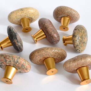 Könnte beinhalten: Acht natürliche Steinknaufgriffe mit goldfarbener Metallbeschläge. Die Knöpfe sind in verschiedenen Brauntönen, Beige und Grau mit einem gesprenkelten Muster.
