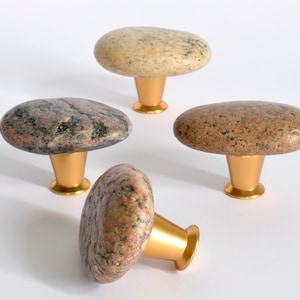 May include: Four natural stone cabinet knobs with gold metal bases. The knobs are shaped like flat, oval stones with a variety of colours and patterns.