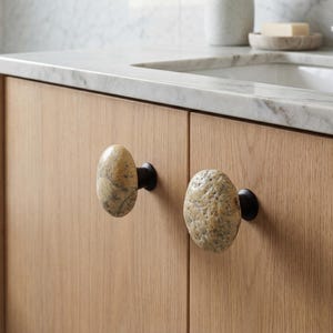 Close-up of unique pebble handles on a chest of drawers, showcasing their polished surfaces and irregular forms. Handmade beach stone drawer pulls add natural charm and tactile interest to nightstands, wardrobes, and storage furniture, combining functional durability with organic design inspired by coastal textures, bringing one-of-a-kind beauty to home interiors.