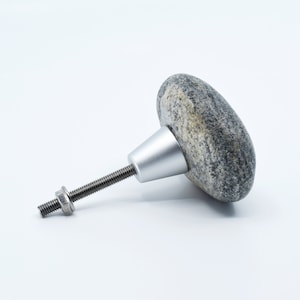 Puede incluir: Un pomo de piedra gris con una base y un tornillo de metal plateado. El pomo tiene forma de piedra plana y redondeada.