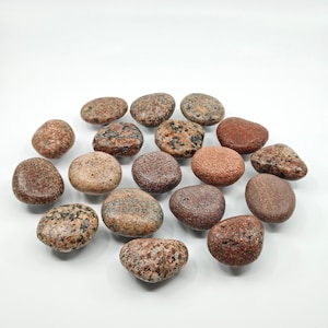 May include: A collection of smooth, rounded river rocks in various shades of brown, red, and grey. The rocks are arranged in a circle on a white background.