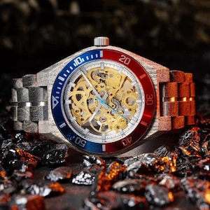 May include: A wooden wristwatch with a skeleton dial, showcasing the intricate inner workings. The watch features a blue and red bezel and a wooden band. The watch is displayed on a dark, textured surface.