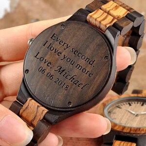 May include: A wooden wristwatch with a dark brown face and band. The watch face has the inscription "Every second, I love you more, Love, Michael 06.06.2018". The watch features a natural wood grain pattern.
