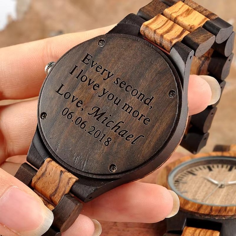 May include: A wooden wristwatch with a dark brown face and band. The watch face has the inscription "Every second, I love you more, Love, Michael 06.06.2018". The watch features a natural wood grain pattern.