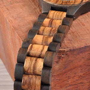 May include: Close-up of a wooden watch with a natural wood grain band. The watch band alternates between dark brown and light brown wood segments. The watch is set against a reddish-brown wooden surface.