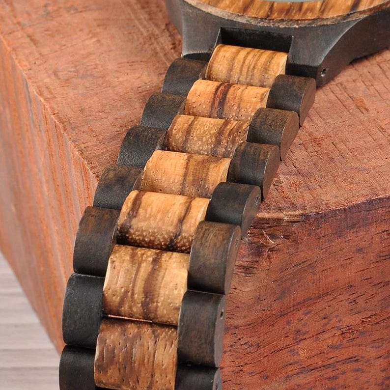 May include: Close-up of a wooden watch with a natural wood grain band. The watch band alternates between dark brown and light brown wood segments. The watch is set against a reddish-brown wooden surface.