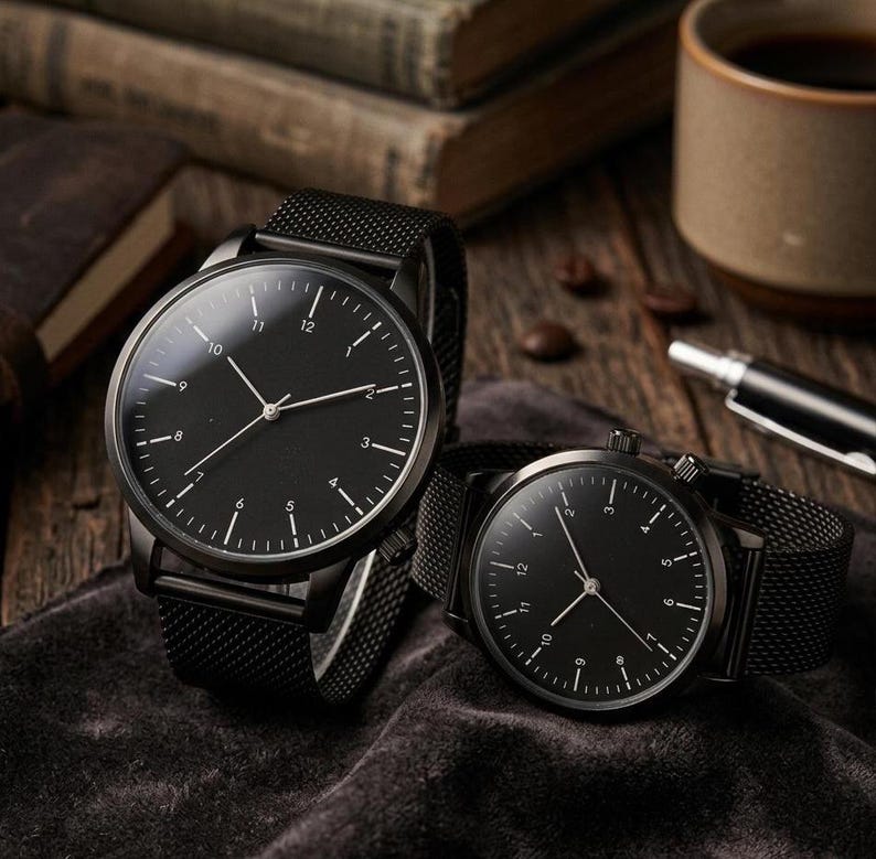 May include: Two black wristwatches with mesh bands are displayed. The watches have black faces with white hour markers and hands. The watches are on a dark gray surface, with a coffee cup and books in the background.