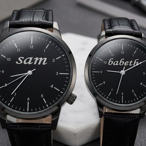 May include: Two black wristwatches with leather straps. One watch face displays the name "sam" in white, the other displays "babeth". Both watches have white hands and hour markers, with a black bezel and case.
