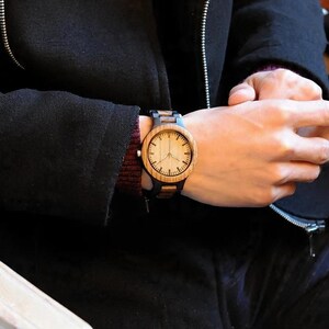 May include: A wooden wristwatch with a light-coloured face and dark brown wooden strap is shown on a wrist. The watch has a classic analog design with a round face and visible hour markers. The watch is a stylish accessory.