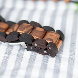 May include: Close-up of a wooden watch strap. The strap is made of rectangular wooden links in shades of brown and black. The watch strap is resting on a white and grey striped surface. The strap has a silver clasp.