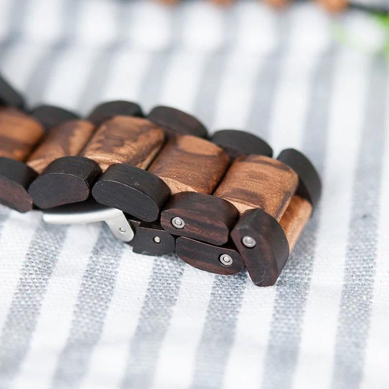 May include: Close-up of a wooden watch strap. The strap is made of rectangular wooden links in shades of brown and black. The watch strap is resting on a white and grey striped surface. The strap has a silver clasp.