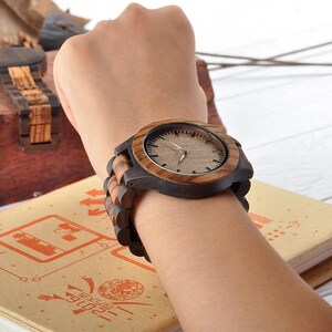 May include: A wooden wristwatch with a dark brown bezel and a light brown wooden watch face. The watch band is made of wooden links. The watch is on a wrist, and the background is blurred.