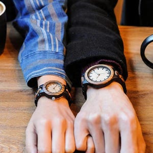 May include: Two wooden wristwatches are shown, each with a light-coloured face and dark strap. The watch faces have a minimalist design with black hands and markers. The watch cases are made of wood, and the straps appear to be dark.
