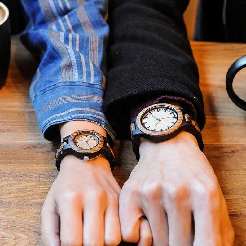 May include: Two wooden wristwatches are shown, each with a light-coloured face and dark strap. The watch faces have a minimalist design with black hands and markers. The watch cases are made of wood, and the straps appear to be dark.