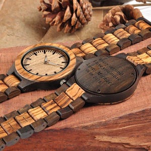 May include: Wooden wristwatch with a light brown face and a dark brown wooden strap. The watch face has black hour markers and hands. The back of the watch is engraved with the words "Every second, I love you more. Love, Michael 06.06.2018."