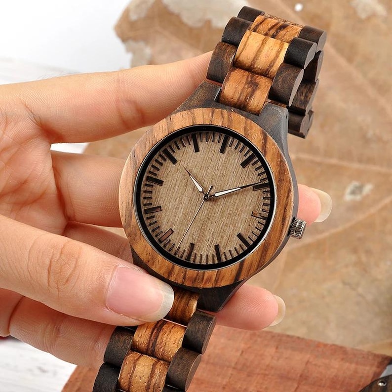 May include: A wooden wristwatch with a light brown face and a dark brown wooden strap. The watch is held in a hand, highlighting the natural wood grain and the watch's design. The watch face has black markings and silver hands.