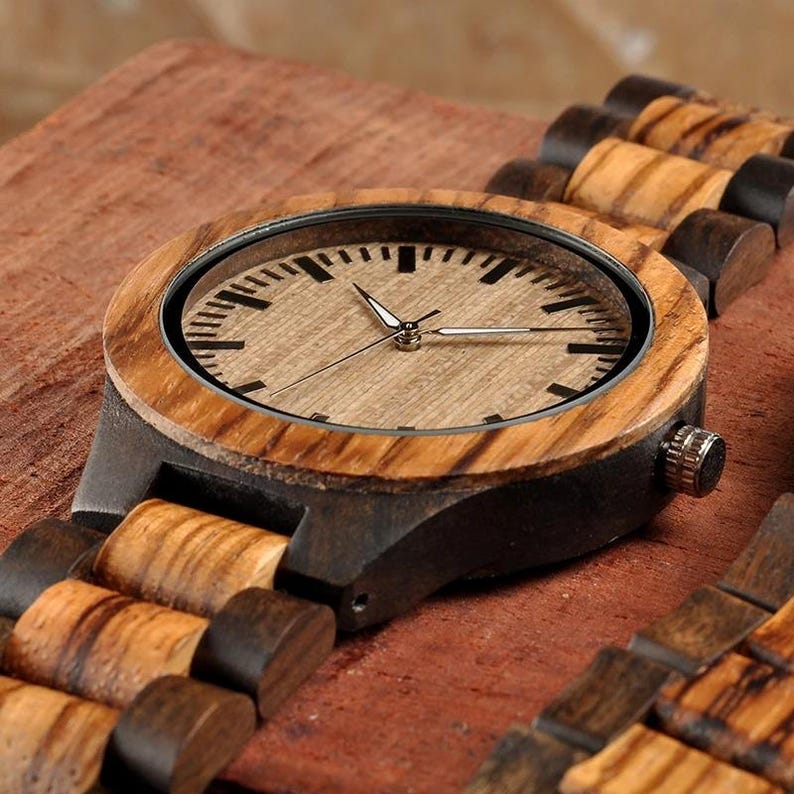 May include: A wooden wristwatch with a light brown face and black hands. The watch has a dark brown case and a wooden strap with alternating light and dark brown segments. The watch is set against a reddish-brown wooden surface.
