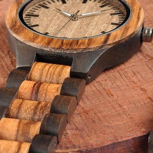 May include: Close-up of a wooden wristwatch with a light brown face and a dark brown wooden band. The watch has silver hands and a silver bezel. The band is made of alternating light and dark brown wooden links.