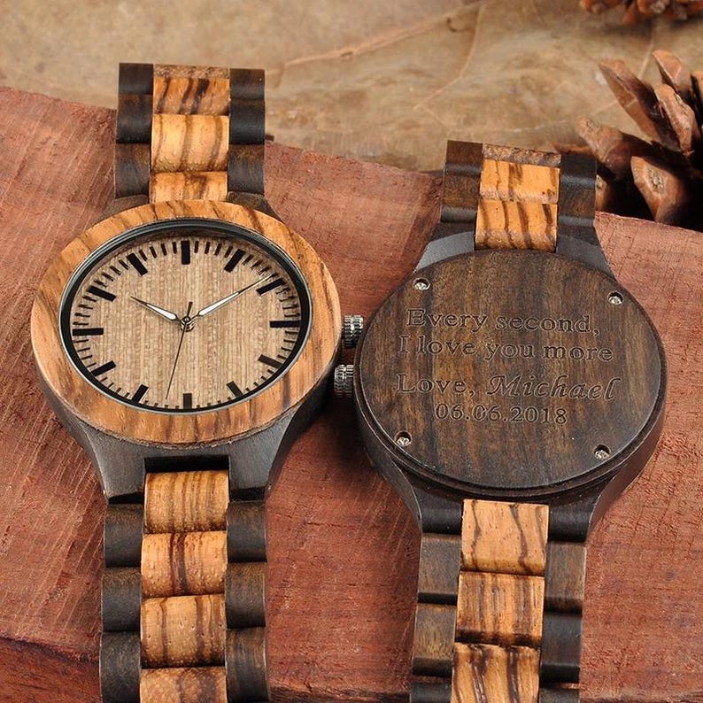 May include: Two wooden watches with a light brown face and dark brown wooden band. The watch face has black hour markers and silver hands. The back of one watch is engraved with the words "Every second, I love you more. Love, Michael 06.06.2018."