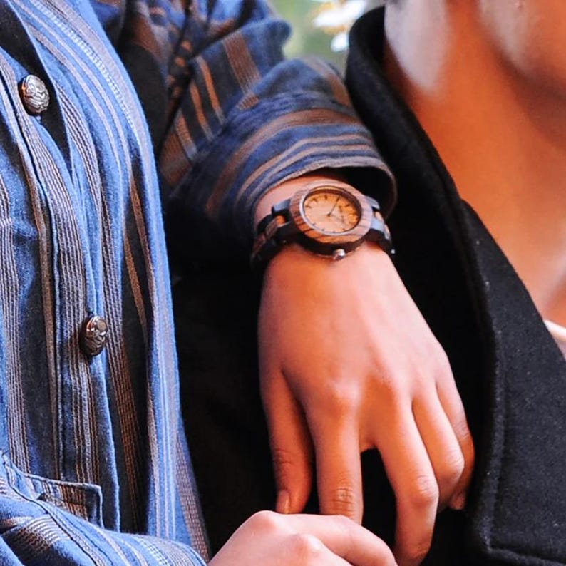 May include: A wooden wristwatch with a light-coloured face and dark wood bezel and strap. The watch is on a wrist, with a blue and brown striped shirt and a black jacket in the background.