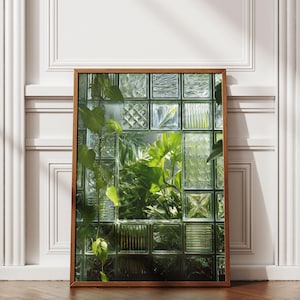 Peut inclure: Une impression encadrée présentant une vue à travers des blocs de verre, révélant des plantes vertes luxuriantes. Le cadre en bois entoure le verre texturé, créant un thème botanique naturel. L'impression est appuyée contre un mur blanc avec des moulures, et le sol est en bois foncé.
