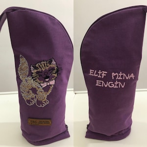 May include: A purple fabric item with a cat embroidery on one side and text on the other. The cat is tan and brown with a pink bow tie. The text says "ELIF MINA ENGIN". A small brown tag is at the bottom.
