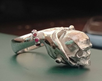 Puede incluir: Un anillo de plata con un diseño de calavera. La calavera está sostenida por una mano con una fila de piedras preciosas rojas en la muñeca.