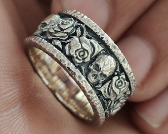 Könnte beinhalten: Ein silberner Ring mit einem Totenkopf- und Rosenmotiv. Der Ring ist breit und hat eine strukturierte Oberfläche.