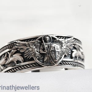 Op de afbeelding: Een zilveren ring met een gegraveerd ontwerp. De ring heeft een centraal embleem van een gevleugelde figuur met een zwaard en schild. De ring heeft een gedetailleerd, gestructureerd patroon en de tekst "badrinathjewellers" is zichtbaar.