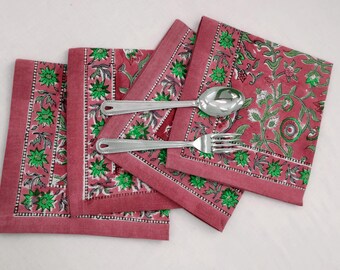 Op de afbeelding: Vier roze katoenen servetten met een groen bloemenpatroon en een witte rand. Een zilveren vork en lepel liggen op de servetten.