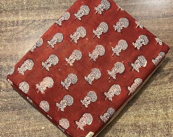 May include: Folded fabric with a repeating floral pattern. The fabric is a deep red color, with white and light pink floral designs. The flowers have a detailed, stylized appearance. The fabric is folded neatly, showing the pattern on the top layer.