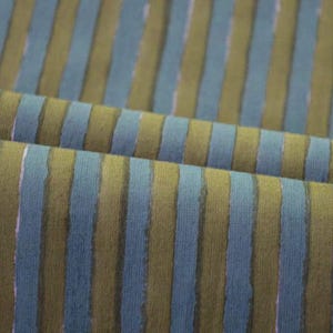 Op de afbeelding: Close-up van een stof met verticale strepen. De strepen wisselen af tussen een gedempt blauwgroen en een gedempt olijfgroen. De stof is gevouwen en toont de textuur en het patroon. De strepen hebben een licht verweerde, handgeschilderde uitstraling.