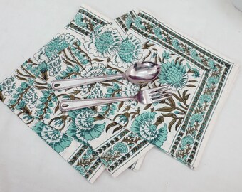 Op de afbeelding: Twee witte katoenen servetten met een teal en bruine bloemenprint. De servetten zijn gevouwen en op elkaar gelegd. Een zilveren vork en lepel liggen op de servetten.