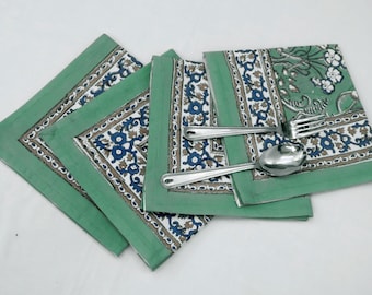 Op de afbeelding: Drie groene katoenen servetten met een wit en blauw bloemenpatroon en een bruine rand. Twee zilveren lepels liggen op de servetten.