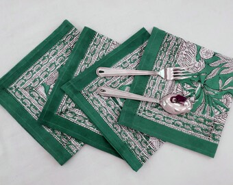 Op de afbeelding: Vier groene katoenen servetten met een witte bloemenprint en een witte rand. De servetten zijn gevouwen en gestapeld. Twee zilveren eetgerei liggen op de servetten.