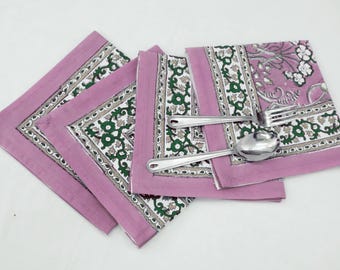 Op de afbeelding: Drie roze katoenen servetten met een groene en witte bloemenprint. De servetten zijn gevouwen en op elkaar gestapeld. Twee zilveren lepels liggen op de servetten.