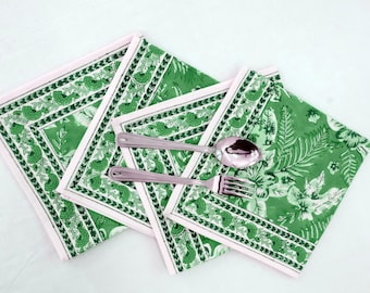 Op de afbeelding: Groene en witte stoffen servetten met een bloemmotief en een roze rand. Een zilveren lepel en vork zijn op de servetten geplaatst. De servetten zijn vierkant.