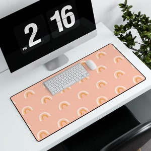 Puede incluir: Una alfombrilla de escritorio rosa con un patrón repetitivo de arcoíris naranja y blanco. Un teclado blanco y un ratón blanco están sobre la alfombrilla.