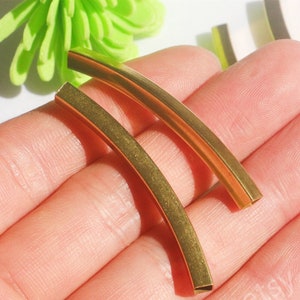 Pode incluir: Dois tubos de metal de cor dourada, curvados em forma de semicírculo, são segurados em uma mão. Os tubos têm um diâmetro de aproximadamente 2,5 cm e um comprimento de 5 cm.