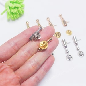 K&ouml;nnte beinhalten: Nahaufnahme eines Sets aus goldenen und silbernen Flaschenverschl&uuml;ssen mit Stecknadel. Die Verschl&uuml;sse sind flaschenf&ouml;rmig und haben eine runde Oberseite. Die Stecknadeln sind an der Unterseite der Verschl&uuml;sse befestigt.