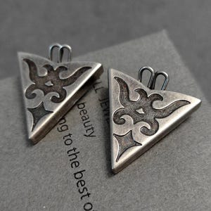 May include: Two triangular silver-toned pendants featuring a dark, recessed filigree pattern with a central stylized motif and a four-pointed star shape at the bottom. Each pendant includes a small, rounded metal loop at the top for attachment.