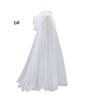 Puede incluir: Una capa con capucha blanca hecha de tela de tul transparente en capas. La prenda presenta una capucha drapeada y una silueta larga y fluida que llega hasta el suelo. El texto b# es visible en la parte superior izquierda de la imagen.