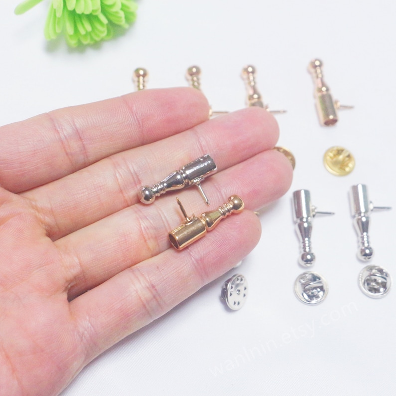 K&ouml;nnte beinhalten: Nahaufnahme von zwei Flaschenverschl&uuml;ssen aus Metall, einer goldfarben und einer silberfarben, mit einer runden Kugel am Ende. Die Verschl&uuml;sse befinden sich auf einer Hand, und im Hintergrund sind weitere Verschl&uuml;sse zu sehen.