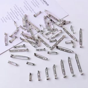 May include: A collection of silver-toned metal brooch pins in various lengths, each featuring a secure clasp mechanism and pre-drilled holes along the flat bar for attachment purposes, scattered across a surface with printed text.