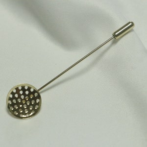 Puede incluir: Un broche de corbata dorado con un diseño redondo perforado. El broche tiene un alfiler largo y delgado.